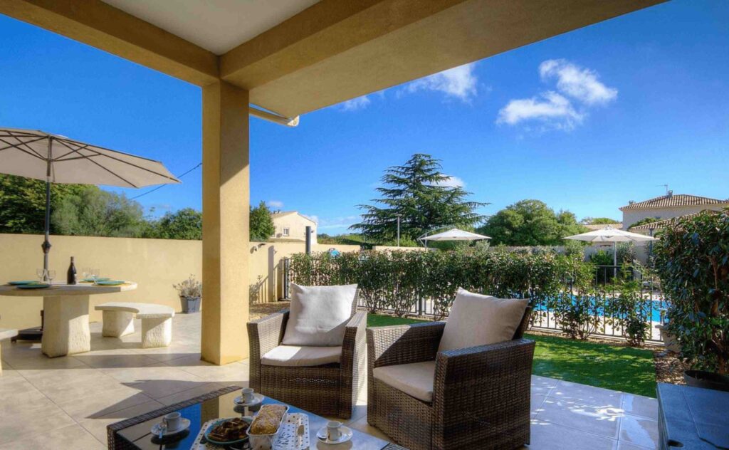 Terrasse couverte avec table et chaises donnant sur une piscine et un jardin.