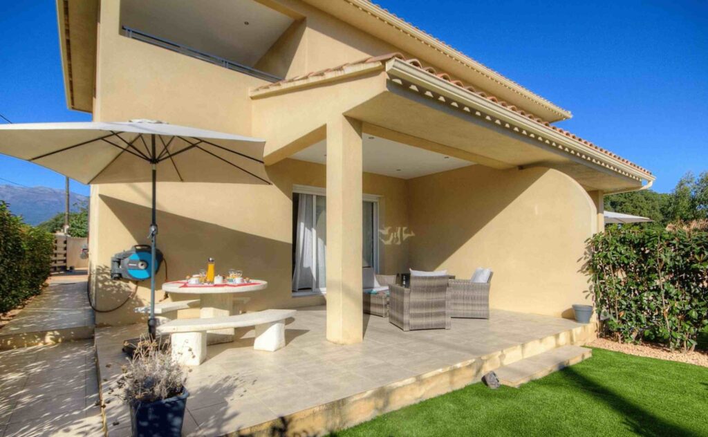 Terrasse avec parasol, table et chaises en plein air devant une maison.