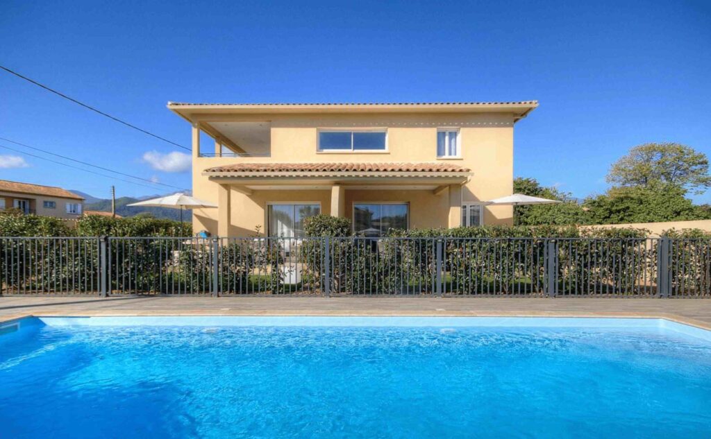 Vue de face de la villa avec une piscine entourée d'une clôture métallique et un ciel bleu clair.