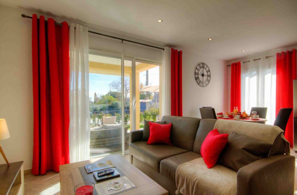 Salon confortable avec canapé gris, coussins rouges, grande baie vitrée donnant sur un jardin.