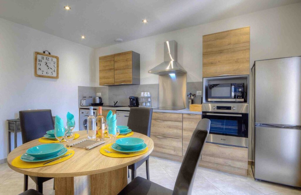 Cuisine moderne avec des éléments en bois, équipée d'électroménagers en acier inoxydable et d'une table à manger ronde décorée avec des assiettes turquoises et des napperons jaunes.